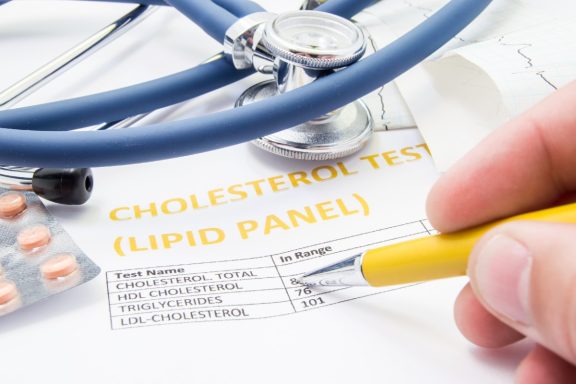 A hand holding a yellow pen points to a cholesterol test report with medication nearby.
